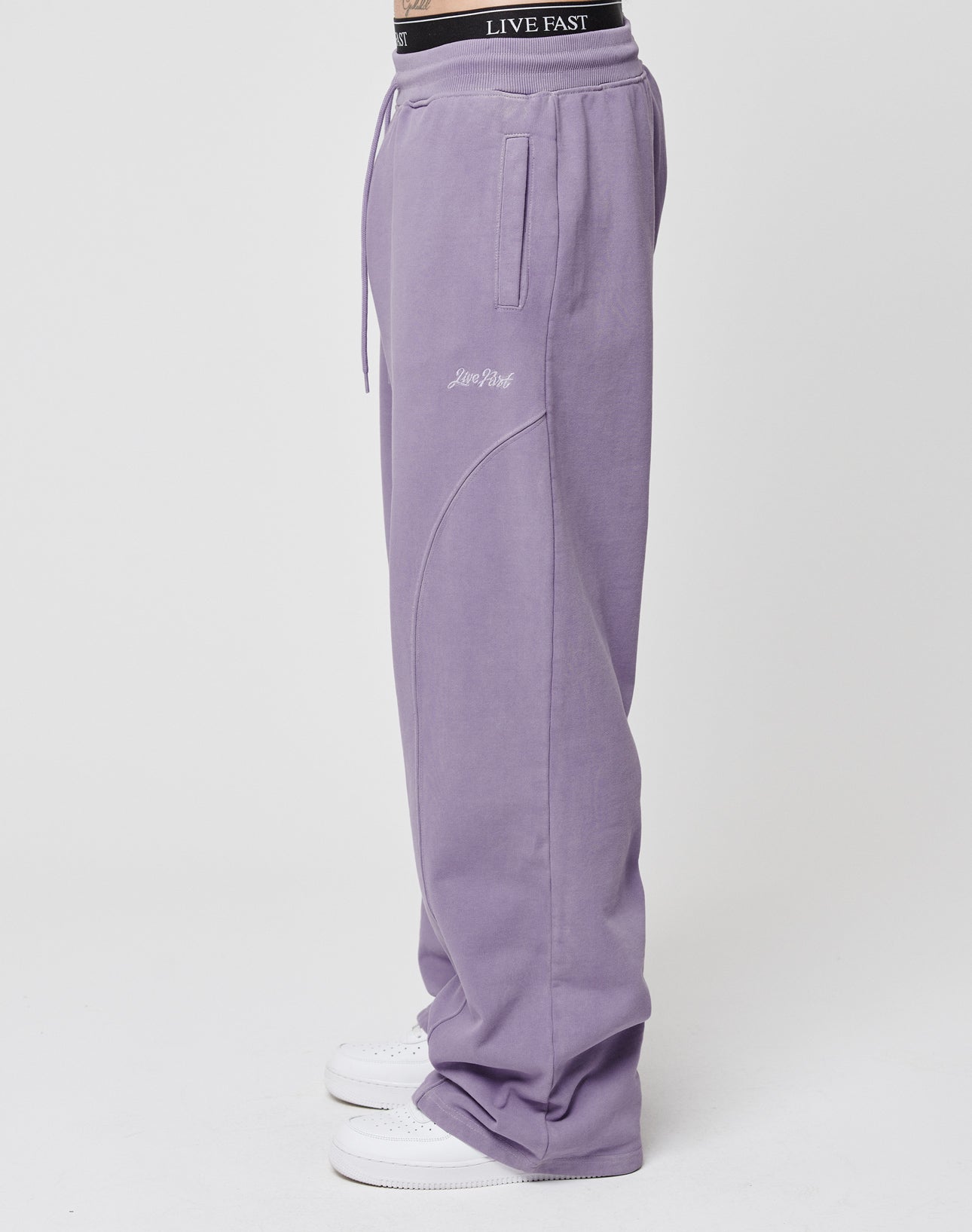 A person stands sideways wearing LIVE FAST DIE YOUNGs Basic Wide Jogger in light purple Baumwolle. The loose-fit pants feature front pockets, seam detailing, an elastic waistband with LIVE FAST, and white sneakers on a plain white background.