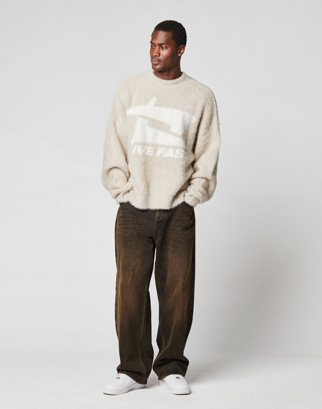 A man stands against a plain light background, wearing LIVE FAST DIE YOUNGs LFDY Baggy Denim brown pants, a fuzzy beige sweater with white graphics and text, and white sneakers. His hands are in his pockets as he looks to the side.