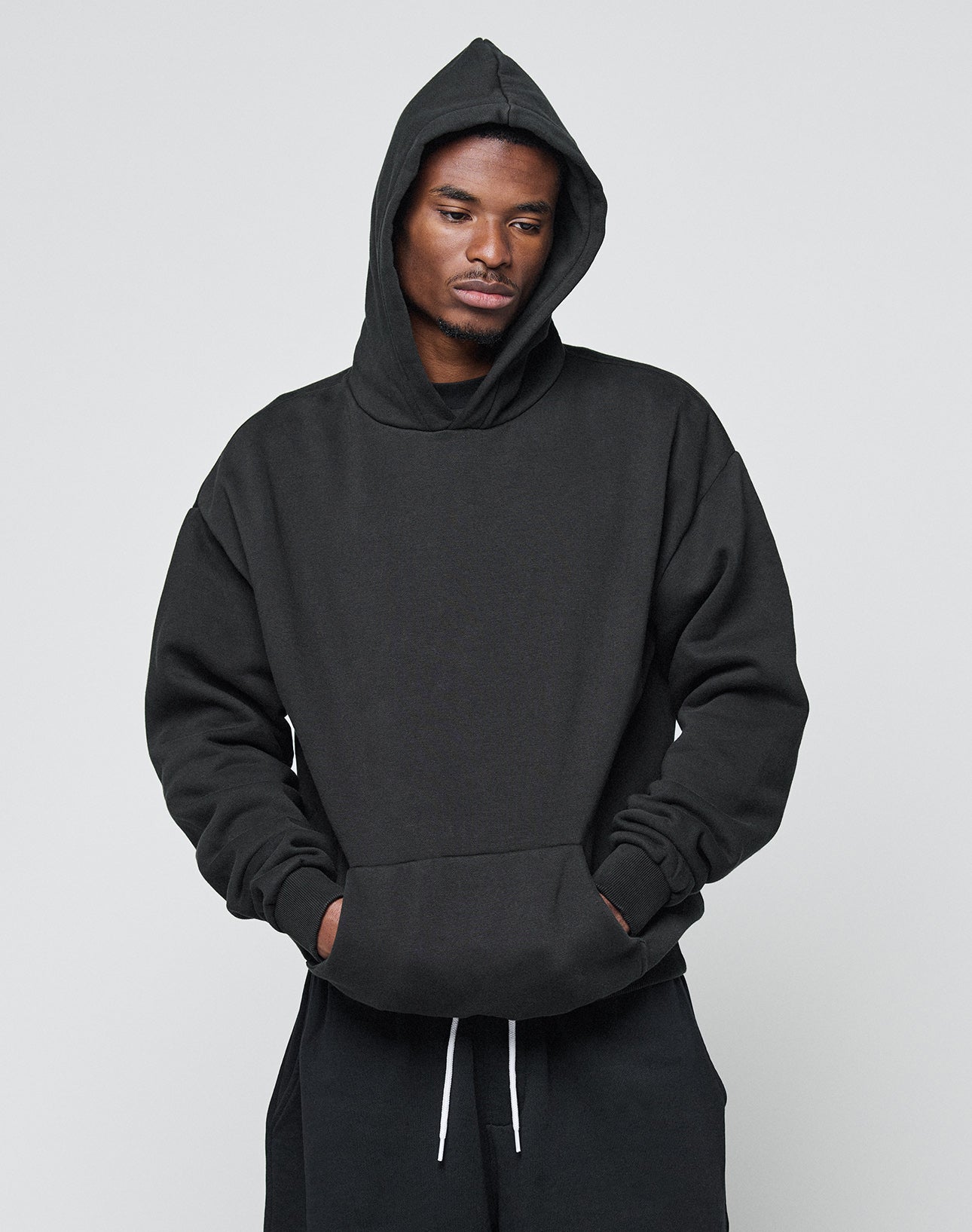 A man wears the LIVE FAST DIE YOUNG Basic Blank Hooded in black, styled oversized with the hood up and paired with black pants, standing against a plain light gray background with his hands in his pockets, looking slightly downward.