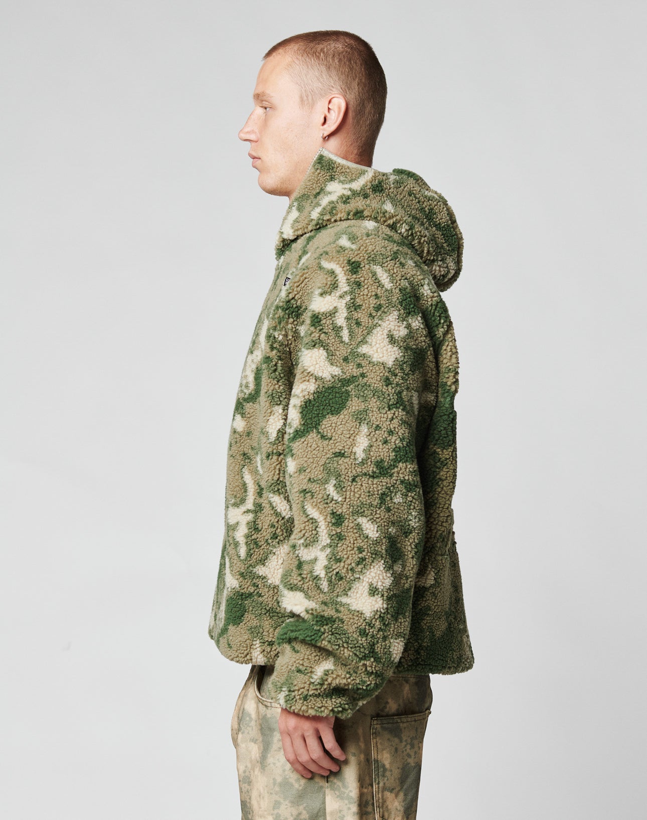 A person stands in profile wearing the LIVE FAST DIE YOUNG Live Fast Teddy Jacket in green and beige camo-patterned fleece with embossed branding, paired with matching pants, against a plain light gray background.