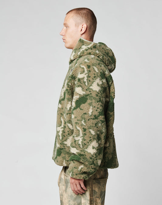 A person stands in profile wearing the LIVE FAST DIE YOUNG Live Fast Teddy Jacket in green and beige camo-patterned fleece with embossed branding, paired with matching pants, against a plain light gray background.