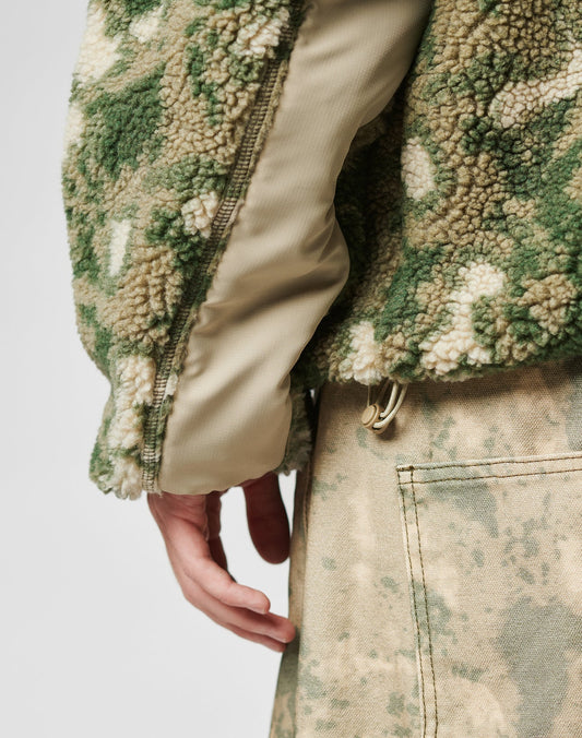 A close-up shows a hand by the side, wearing the LIVE FAST DIE YOUNG Live Fast Teddy Jacket in green and beige camo with a beige panel and embossed branding, paired with light camo pants featuring a visible pocket.