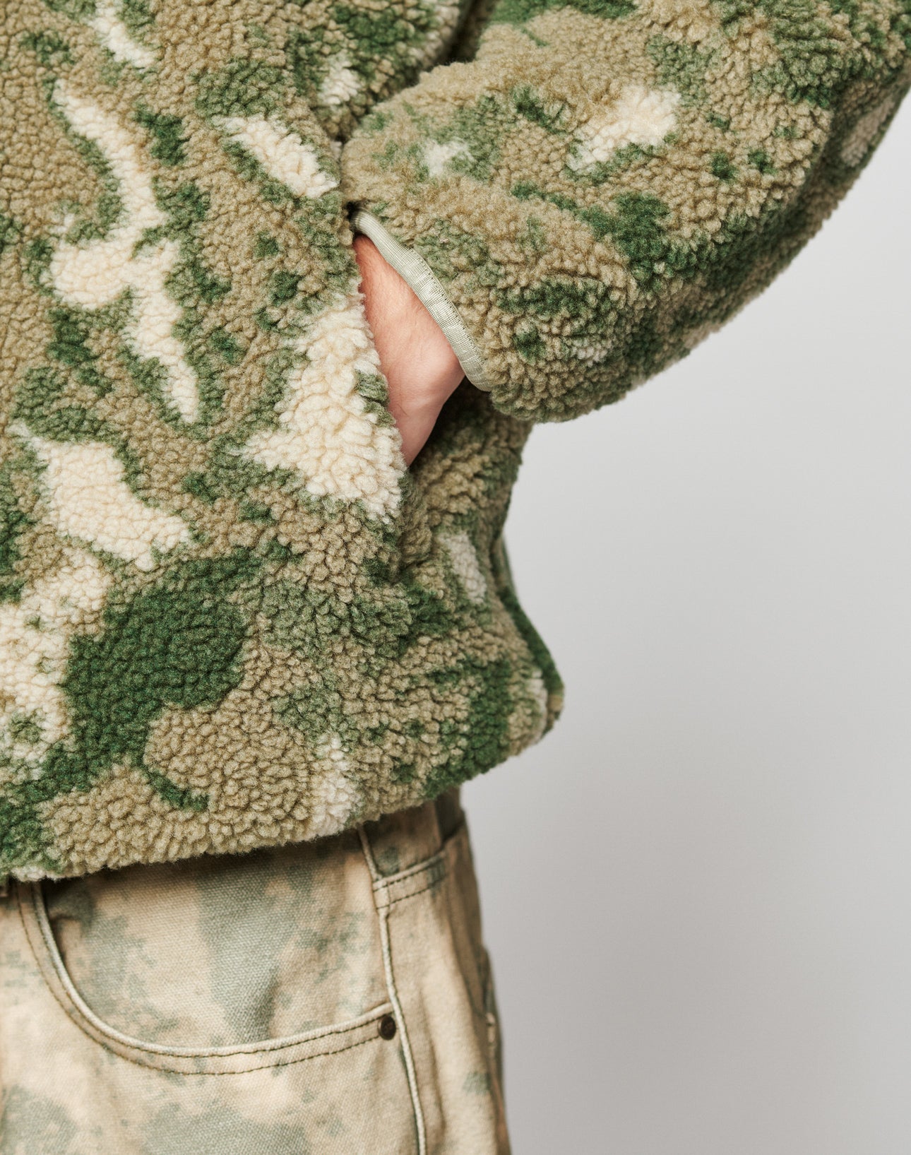 A person with their hand in the pocket of a green and beige camouflage Live Fast Teddy Jacket by LIVE FAST DIE YOUNG, featuring embossed branding, wears matching camo pants against a plain light background.