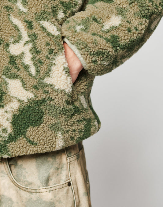 A person with their hand in the pocket of a green and beige camouflage Live Fast Teddy Jacket by LIVE FAST DIE YOUNG, featuring embossed branding, wears matching camo pants against a plain light background.