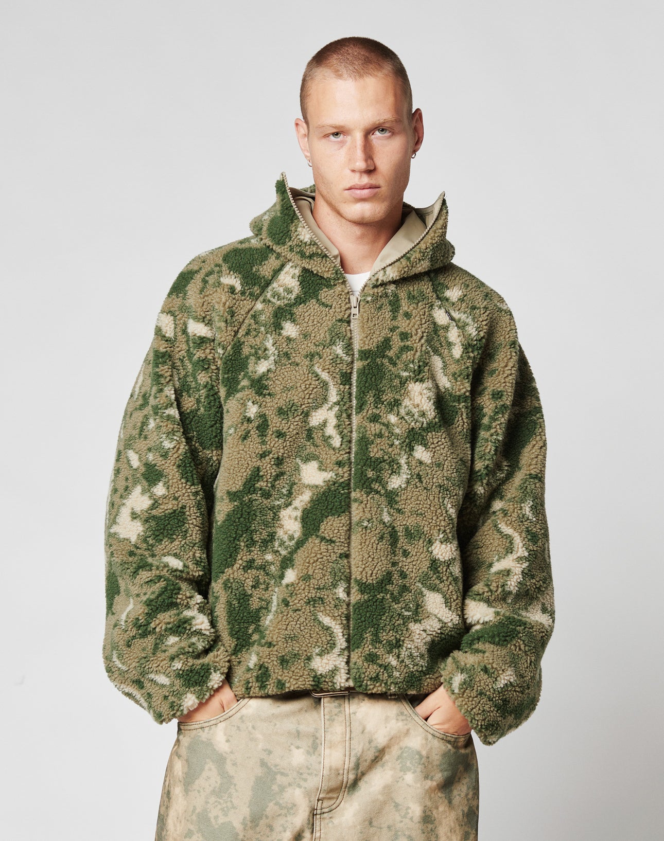 A person models the LIVE FAST DIE YOUNG Live Fast Teddy Jacket—a green and beige camo oversize fleece hoodie with zip-up front and visor hood—standing against a plain light gray background, hands in pockets.