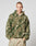 A person models the LIVE FAST DIE YOUNG Live Fast Teddy Jacket—a green and beige camo oversize fleece hoodie with zip-up front and visor hood—standing against a plain light gray background, hands in pockets.