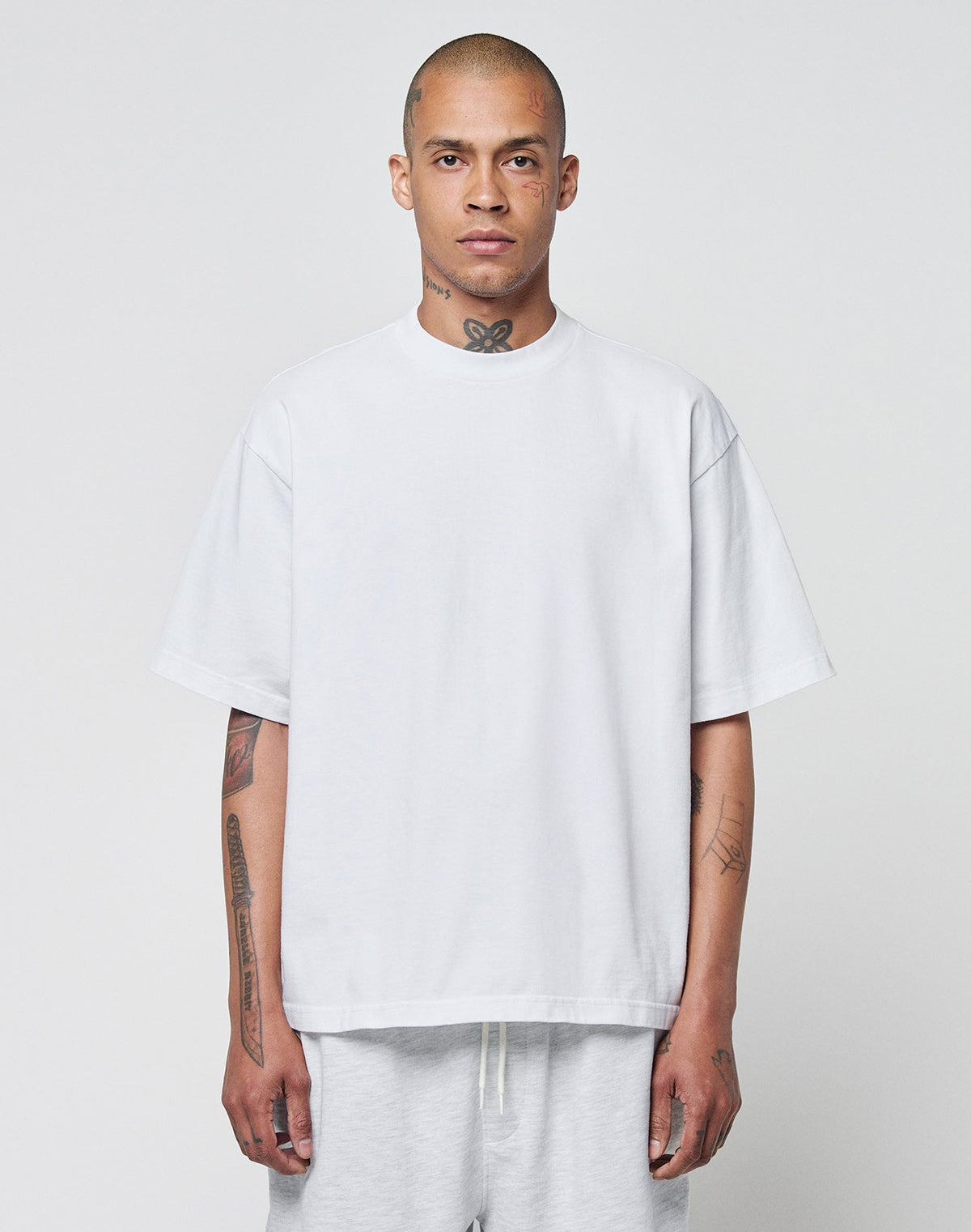 A man with a shaved head and tattoos wears the LIVE FAST DIE YOUNG Basic Heavy Tee, an oversize fit white t-shirt made from 100% cotton, paired with light pants. He stands against a plain light gray background, looking at the camera. | model