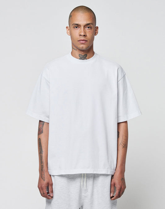 A man with a shaved head and tattoos wears the LIVE FAST DIE YOUNG Basic Heavy Tee, an oversize fit white t-shirt made from 100% cotton, paired with light pants. He stands against a plain light gray background, looking at the camera. | model