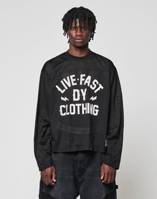 A young man with short dreadlocks models the LFDY Flash Jersey by LIVE FAST DIE YOUNG, an oversize black long-sleeve shirt with a bold white front print, standing against a plain light gray background. | model