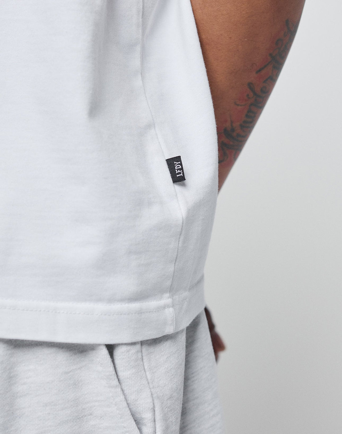 Close-up of a person in the LIVE FAST DIE YOUNG Basic Heavy Tee, an oversize fit white T-shirt made from 100% cotton, paired with light gray pants. A tattoo is on the arm, and a black fabric tag with white text is at the hem.
