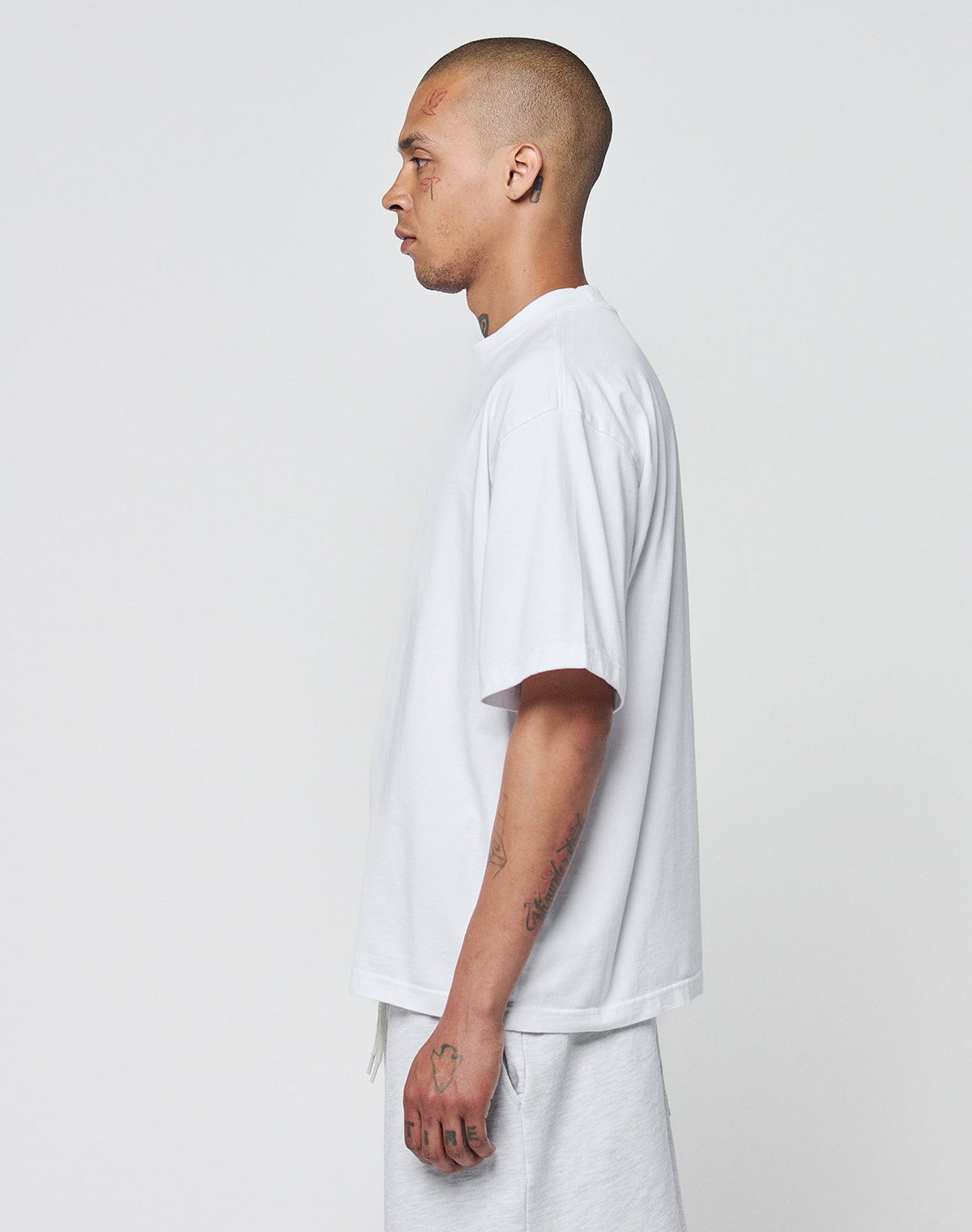 A tattooed, shaved-head person stands in profile against a plain background, wearing the LIVE FAST DIE YOUNG Basic Heavy Tee in oversized white 100% Baumwolle and matching white shorts.