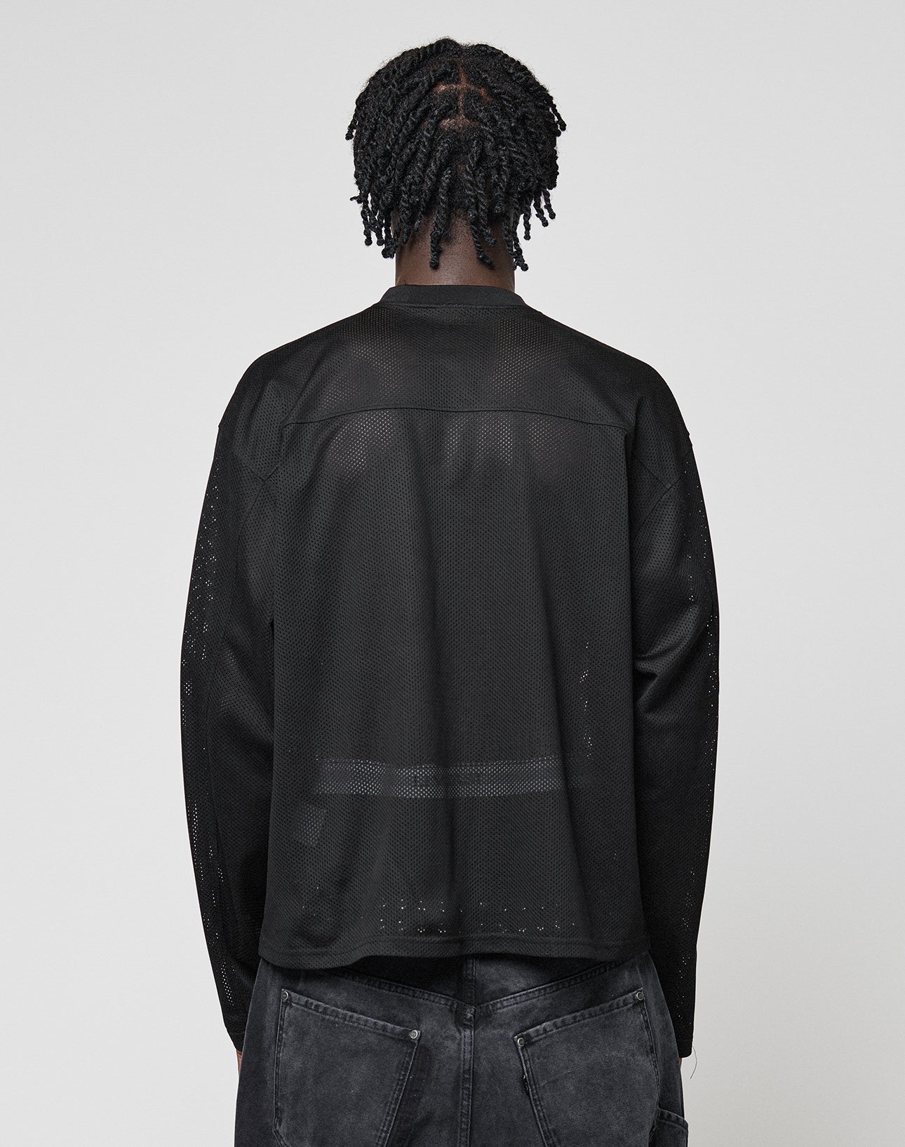 A person with short, twisted braids is seen from behind wearing the LFDY Flash Jersey by LIVE FAST DIE YOUNG—an oversize-fit, long-sleeve black mesh top—paired with faded black jeans against a plain light gray background.