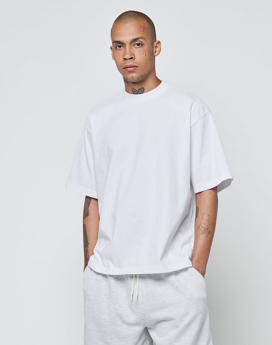 A tattooed man stands against a plain background, wearing the LIVE FAST DIE YOUNG Basic Heavy Tee in oversize fit made from 100% Baumwolle and light-colored pants, with hands in his pockets and a neutral expression.