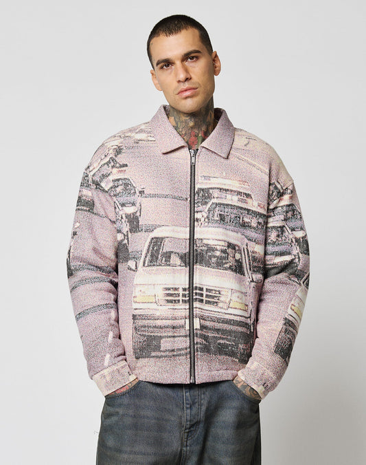 A person with short hair and neck tattoos wears the LIVE FAST DIE YOUNG LFDY Chase Jacket, an oversize transitional jacket with a car traffic graphic print. The plain light background and persons neutral expression complete the look.