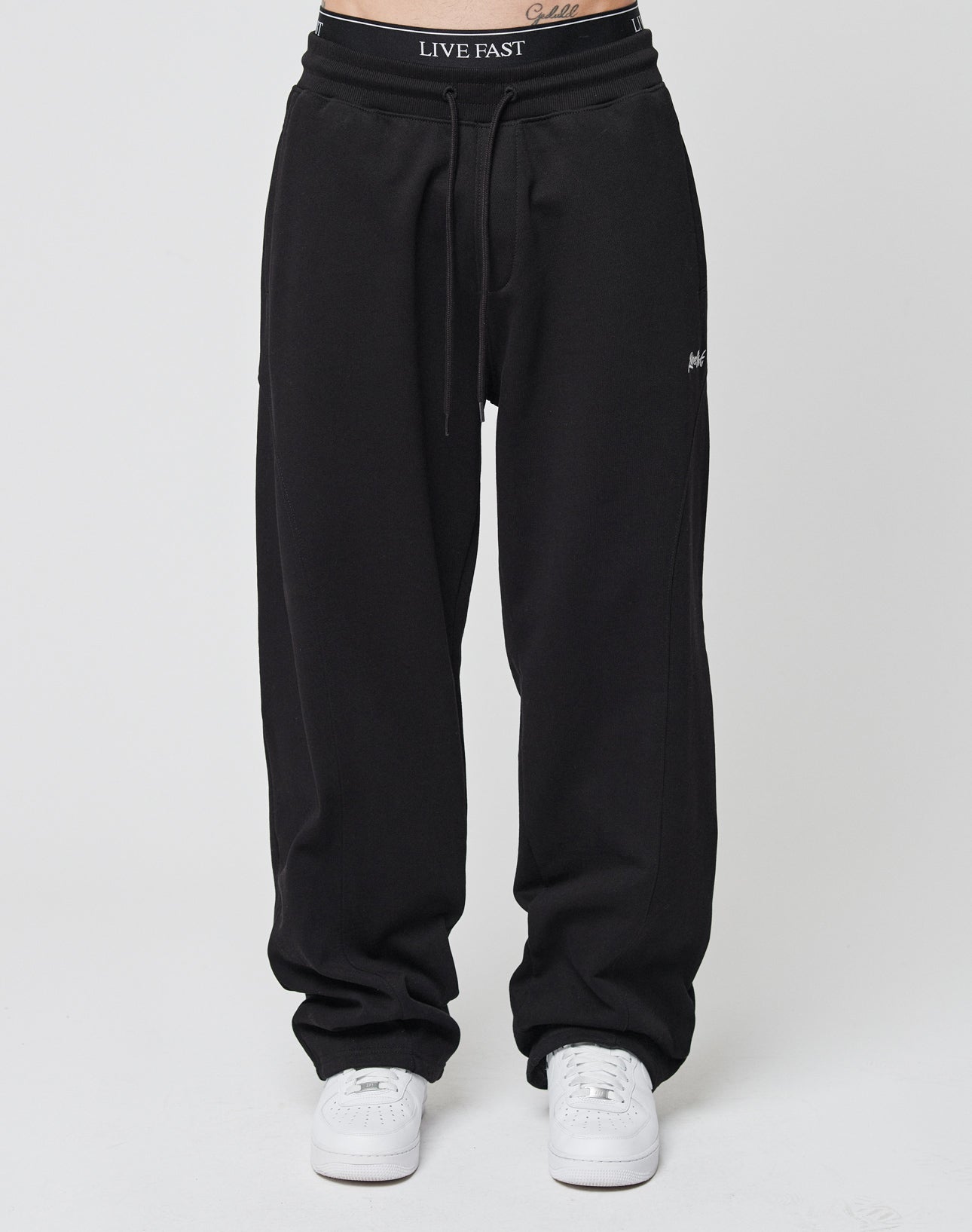 A person wears the Basic Wide Jogger by LIVE FAST DIE YOUNG—loose black sweatpants with a wide fit and elastic waistband featuring LIVE FAST—paired with white sneakers. The photo is chest-down, showing a small abdomen tattoo on a plain background.
