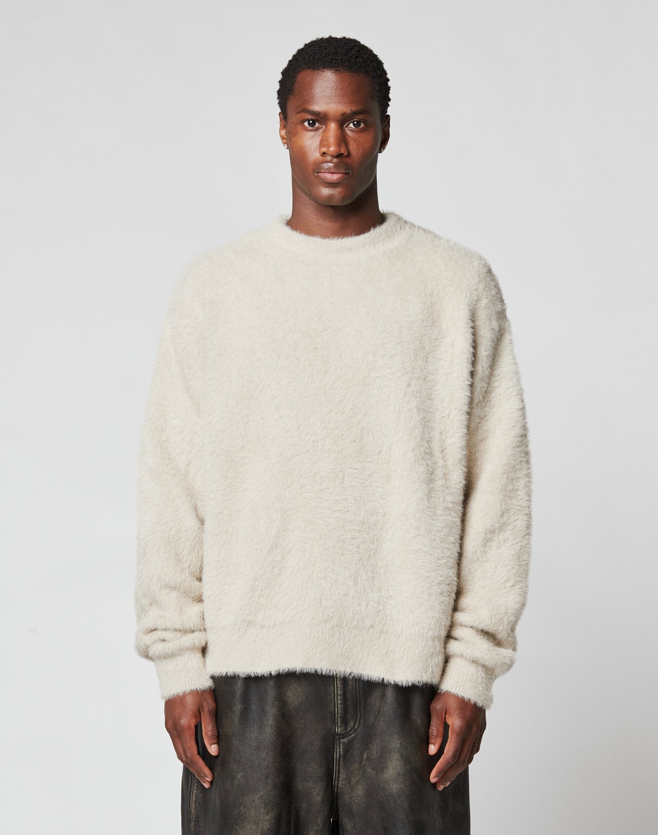 A person stands facing forward against a plain light background, wearing the cream-colored LIVE FAST DIE YOUNG LFDY Praying Fuzzy Sweater with an oversize fit and dark, loose-fitting pants.