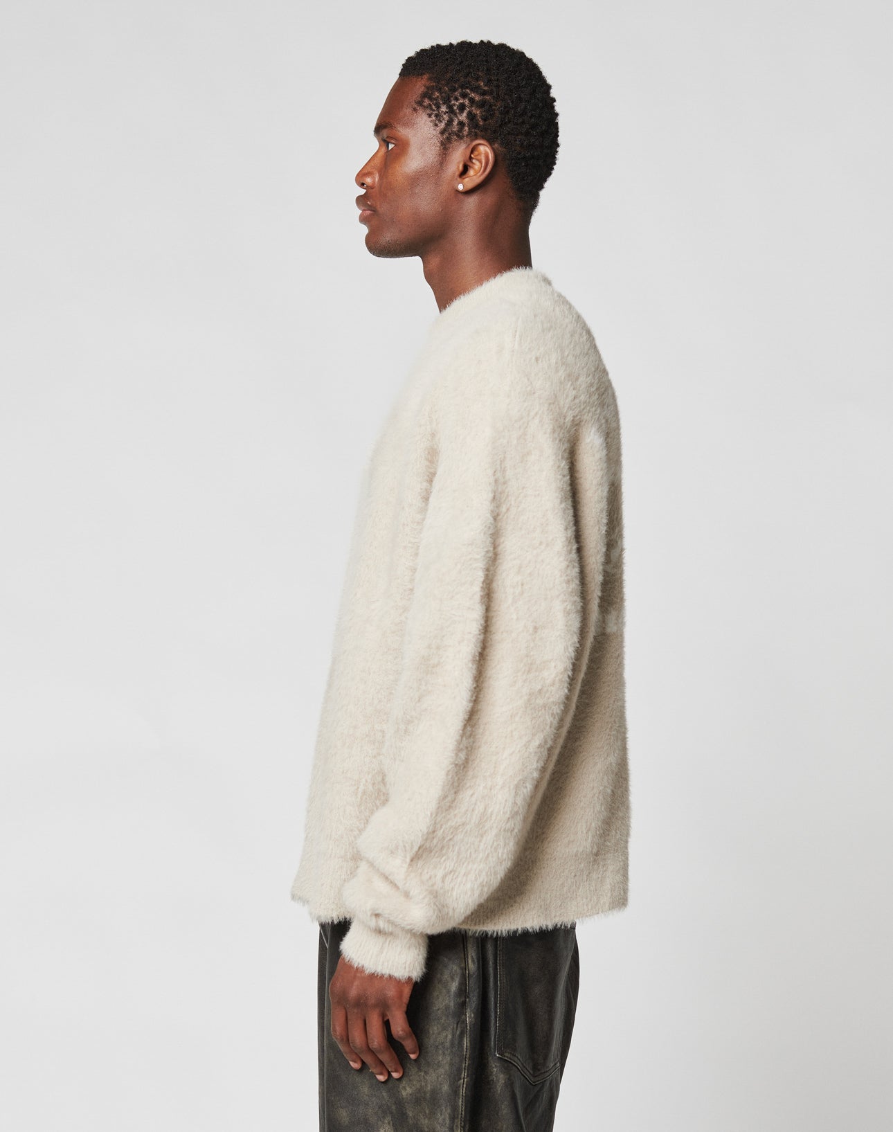 A person stands in profile against a plain background, wearing the off-white LIVE FAST DIE YOUNG LFDY Praying Fuzzy Sweater with Rundhals neckline and Oversize Fit, paired with dark pants.