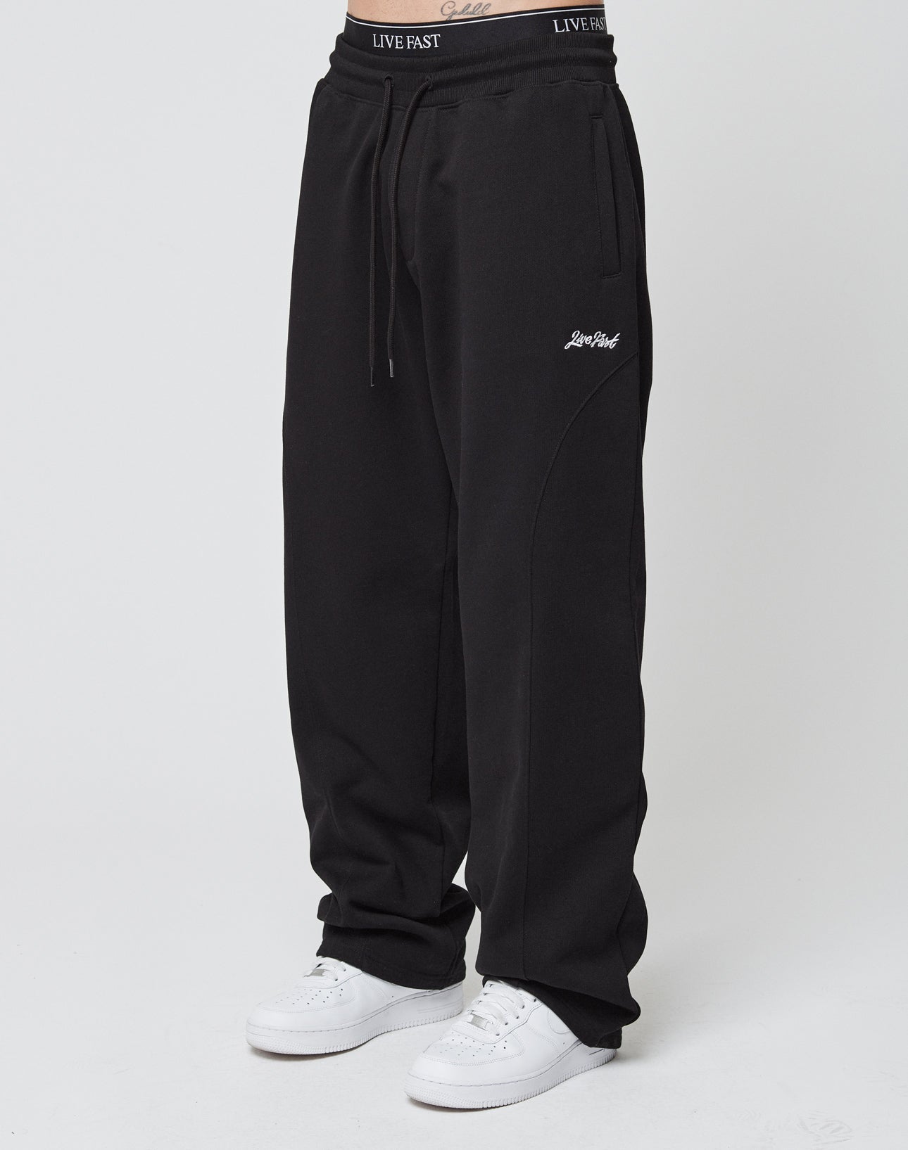 A person wears Basic Wide Jogger sweatpants from LIVE FAST DIE YOUNG, featuring a LIVE FAST logo on the waistband and left thigh. They pair them with white sneakers against a plain, light background.