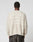 A person with short curly hair is seen from behind wearing the LIVE FAST DIE YOUNG LFDY Praying Fuzzy Sweater—an oversize beige sweater with intarsia logo, white graphics, a cross, and hands—paired with dark pants.