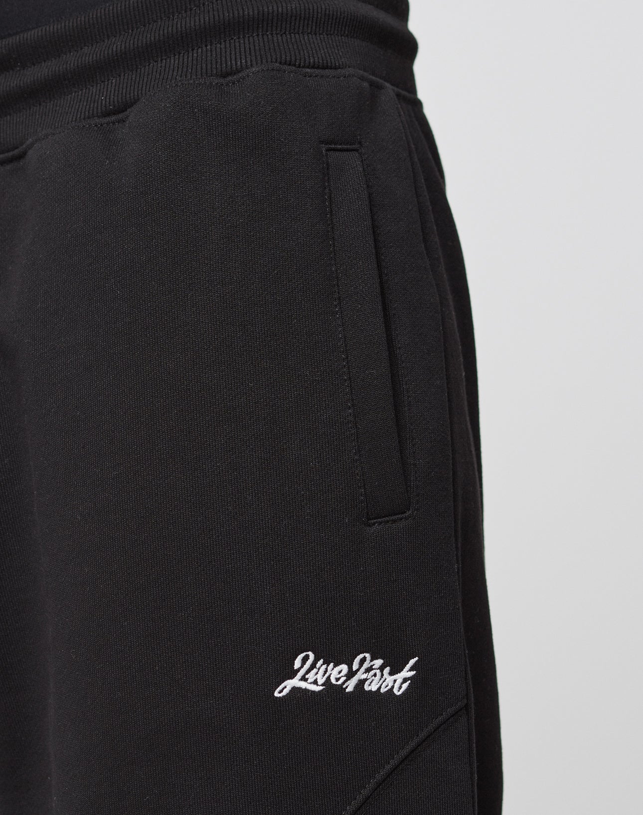 Close-up of the Basic Wide Jogger by LIVE FAST DIE YOUNG: black, wide-fit sweatpants with an elastic waistband, side pockets, and white Live Fast embroidery on the thigh. Made from soft cotton for all-day comfort.