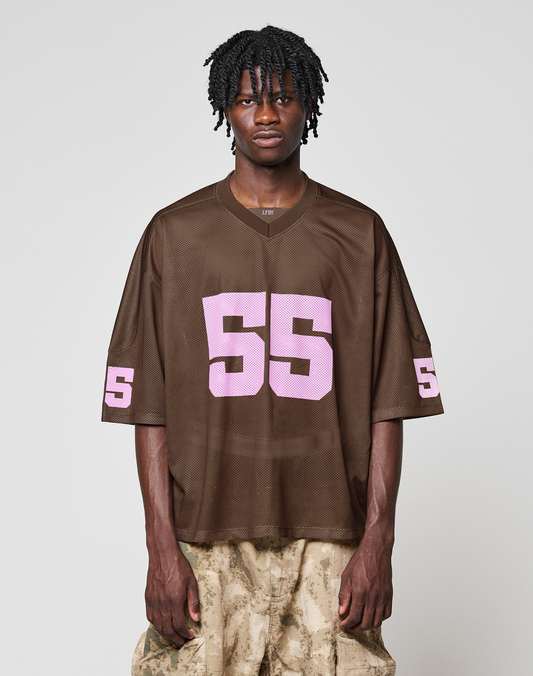 A person with short, twisted hair wears a loose brown LFDY Mesh Jersey by LIVE FAST DIE YOUNG with a pink 55 and V-neck, paired with light brown camo pants, standing against a plain light background. | model