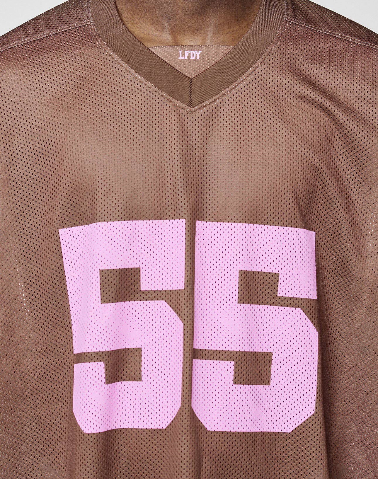 A person wears the LFDY Mesh Jersey by LIVE FAST DIE YOUNG: a brown, oversized mesh top with a V-neck, large pink 55 on the front, and LFDY lettering near the collar.