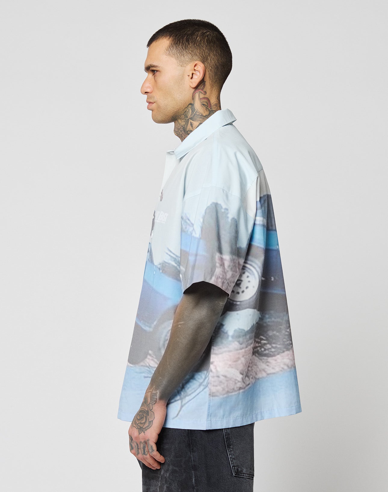 A man with short hair and tattoos stands in profile against a plain light background, wearing a loose, oversize LIVE FAST DIE YOUNG LFDY Crash Kid Shirt with an abstract car and landscape print, paired with dark pants.