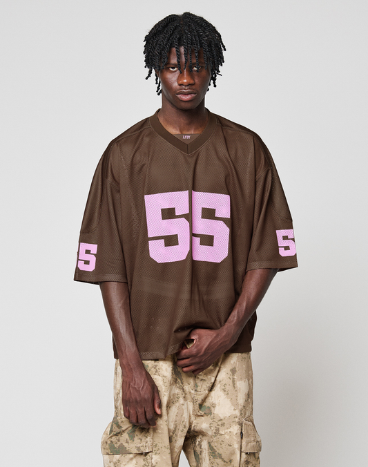 A person stands against a plain background wearing an oversize brown LIVE FAST DIE YOUNG LFDY Mesh Jersey with a V-neck and pink 55, paired with light tan camo-print polyester pants.
