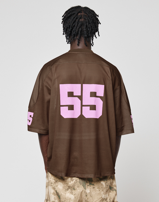 A person with short, twisted hair is shown from behind wearing the oversize fit LFDY Mesh Jersey by LIVE FAST DIE YOUNG in brown polyester mesh, featuring a large pink 55 on the back and left sleeve, paired with tan camouflage pants.