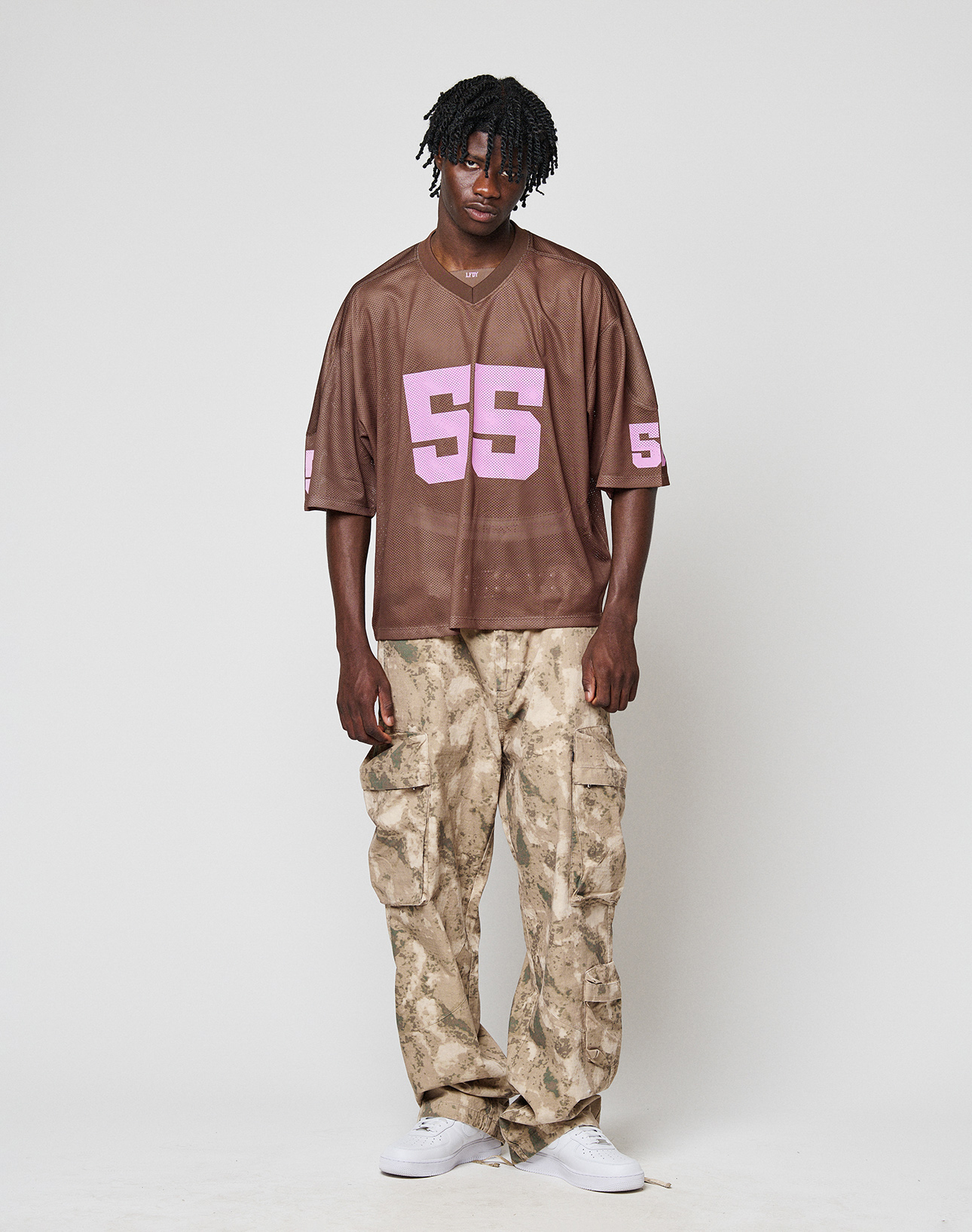 A person with short dreadlocks and a relaxed posture stands against a plain background, wearing a loose brown sports jersey with pink “55,” LIVE FAST DIE YOUNG LFDY Cargo beige camouflage wide fit cargo pants, and white sneakers.