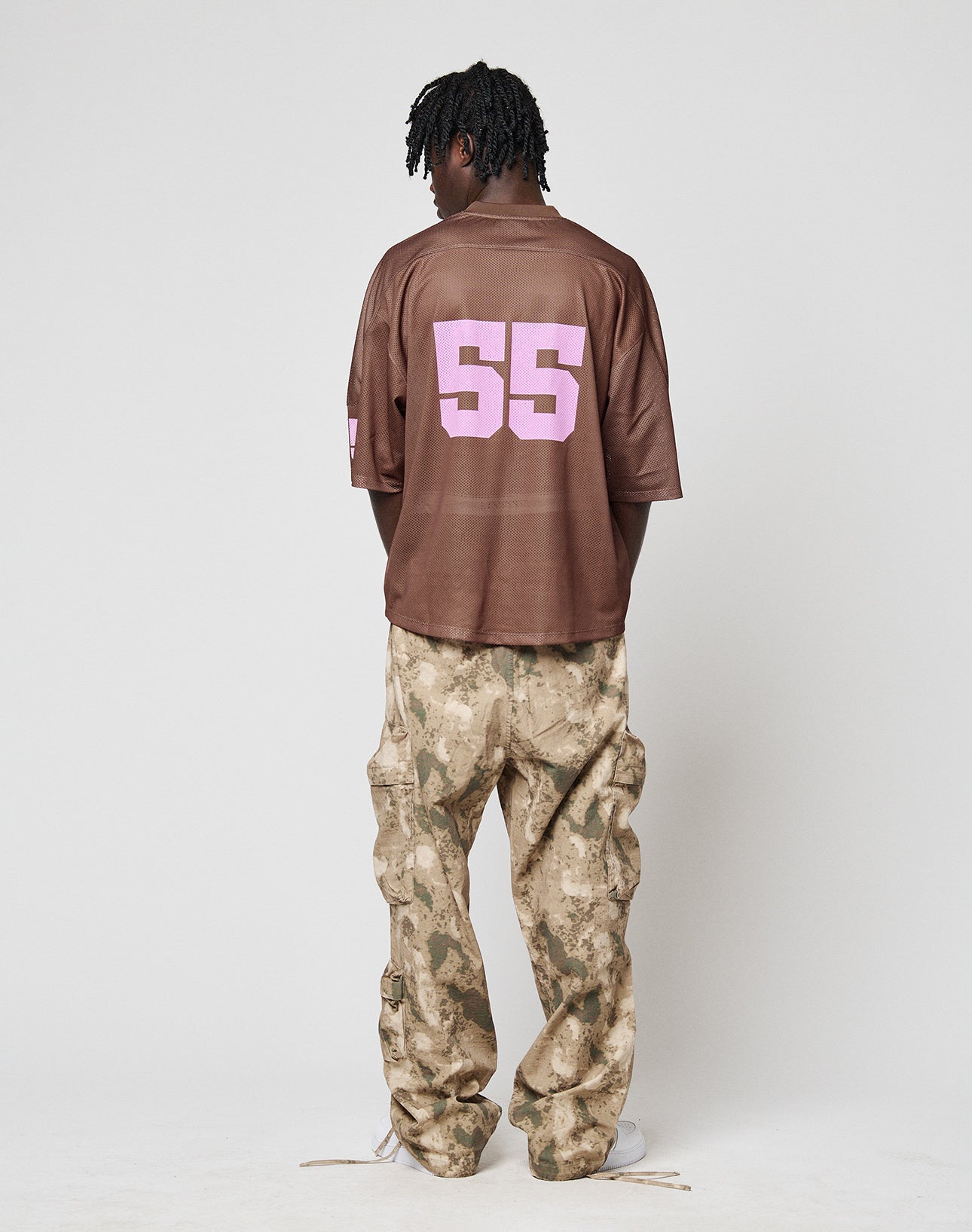 A person faces away from the camera, wearing an oversize LFDY Mesh Jersey by LIVE FAST DIE YOUNG with a large pink 55 on the back and loose tan camo pants. The background is plain and light-colored.