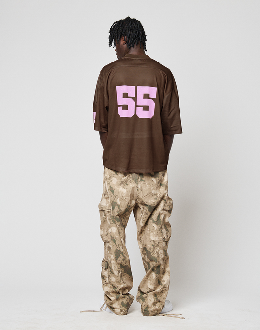 A person faces away from the camera wearing a brown LFDY Mesh Jersey by LIVE FAST DIE YOUNG, featuring a pink 55 on the back, paired with loose tan camo cargo pants. The oversized fit and plain white backdrop complete the look.