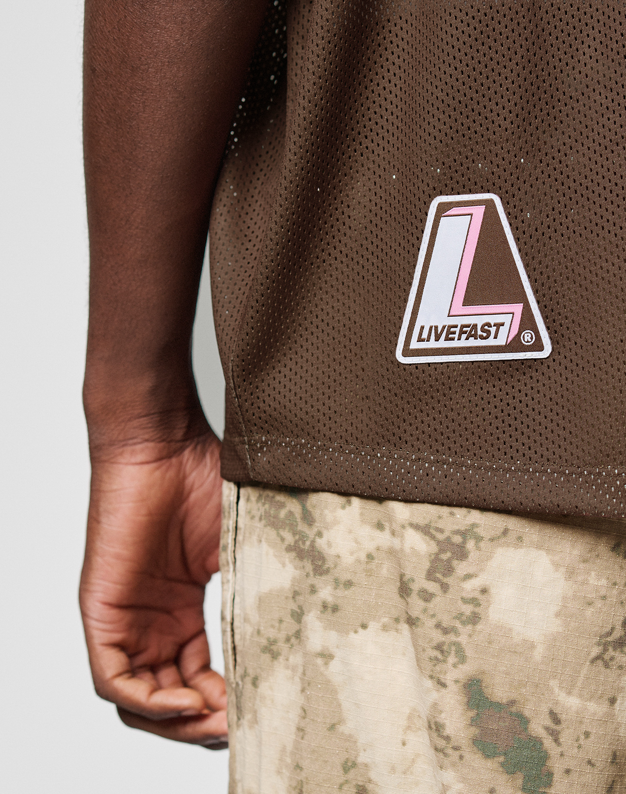 A close-up of a person wearing the LIVE FAST DIE YOUNG LFDY Mesh Jersey, an oversize brown mesh shirt with a pink and white L LIVEFAST patch, paired with camouflage pants. Their left arm hangs relaxed by their side.