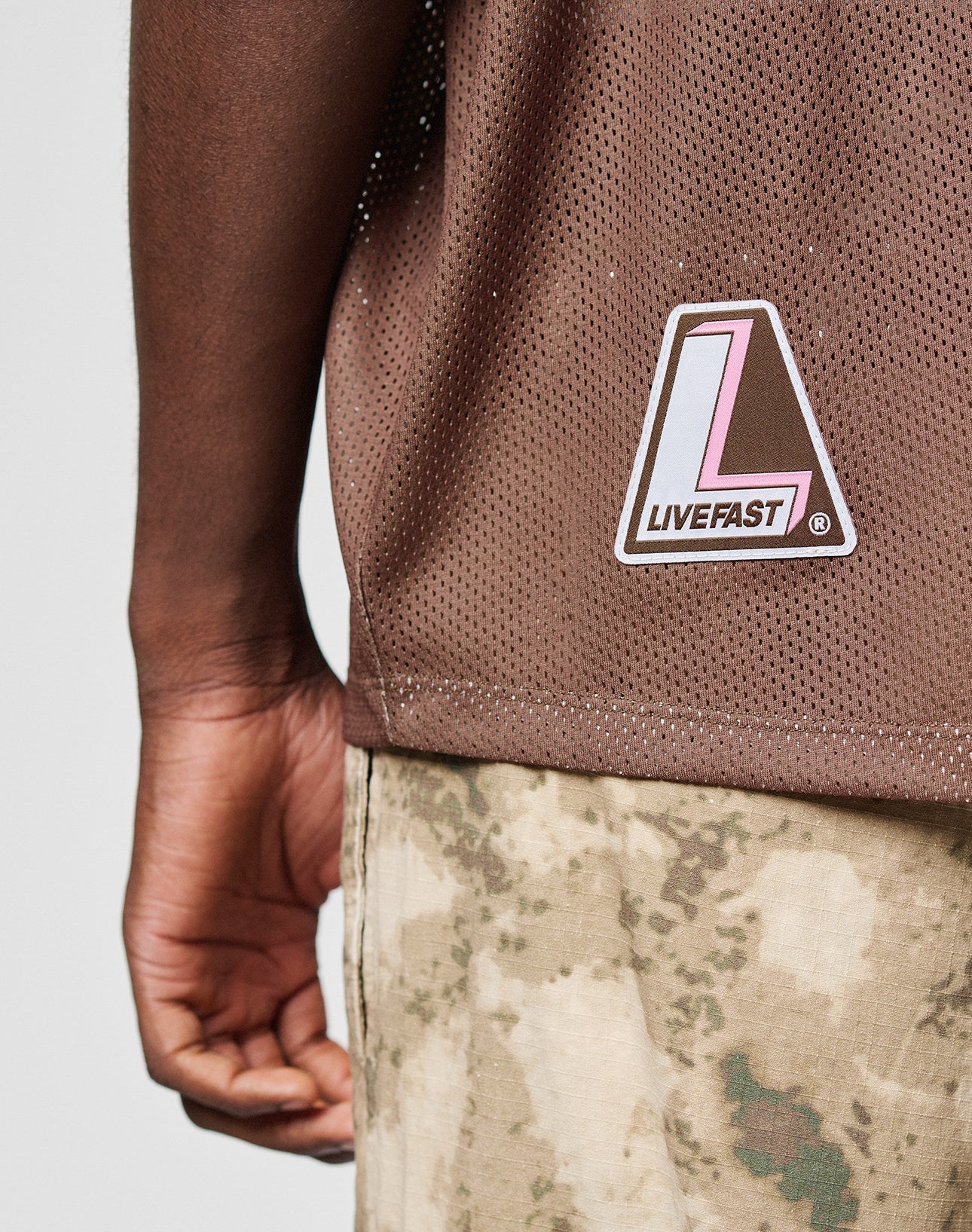 A person is seen from the back wearing the LIVE FAST DIE YOUNG LFDY Mesh Jersey—a brown, oversized mesh shirt with a LIVEFAST logo patch—paired with camouflage pants and one arm visible.