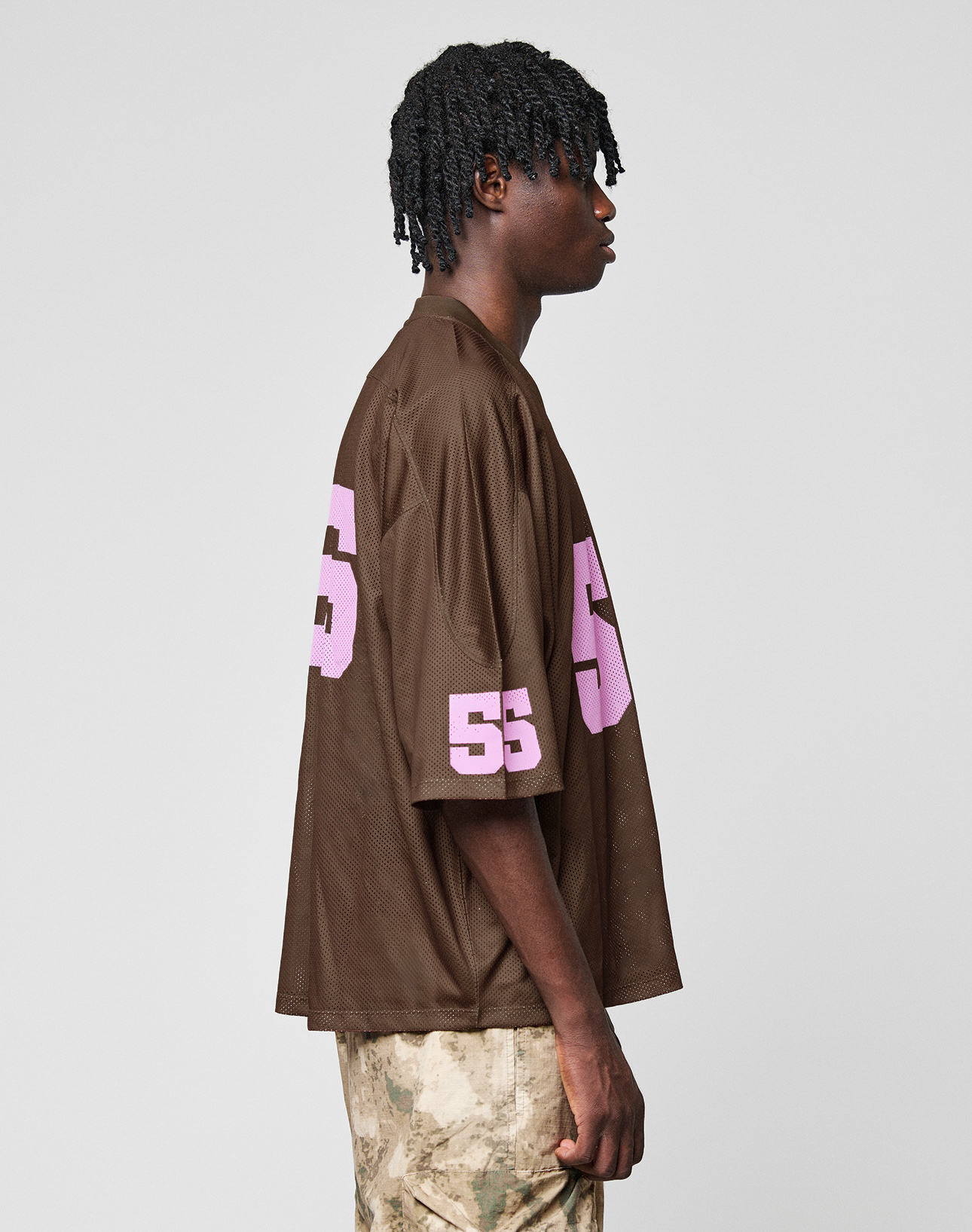 A person in profile wears the LIVE FAST DIE YOUNG LFDY Mesh Jersey—an oversized brown mesh top with a V-neck and pink 55 on sleeve and back—paired with camo pants, set against a plain light gray background.