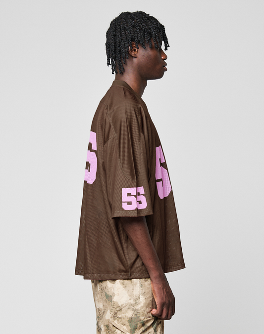 A person in profile wears the LIVE FAST DIE YOUNG LFDY Mesh Jersey—an oversized brown mesh top with a V-neck and pink 55 on sleeve and back—paired with camo pants, set against a plain light gray background.
