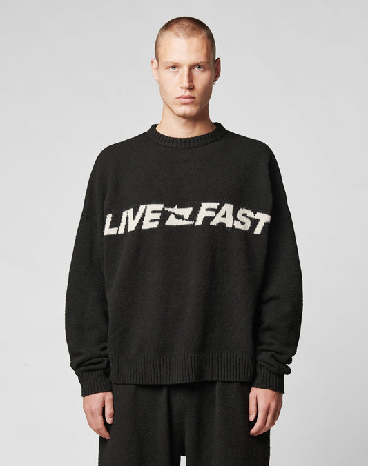A person with short hair models the LIVE FAST DIE YOUNG Live Fast Boucle Sweater—an oversize black polyester piece with LIVE FAST in bold white across the chest—against a plain light gray background.