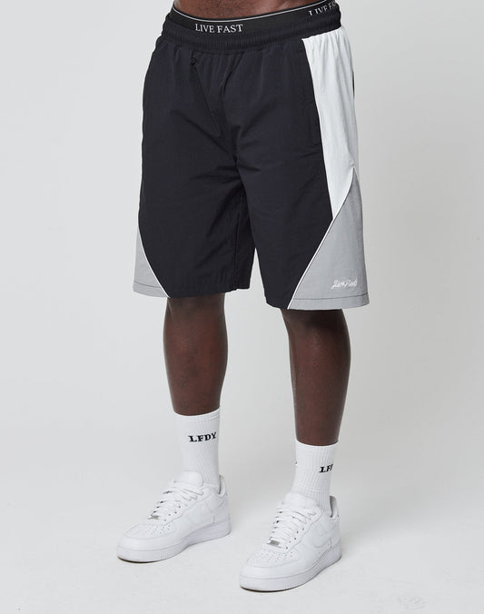 A person in black, gray, and white LIVE FAST DIE YOUNG Basic Nylon Trackshorts with elastic waistband and logo embroidery, white LFDY socks, and white sneakers stands against a plain white background.