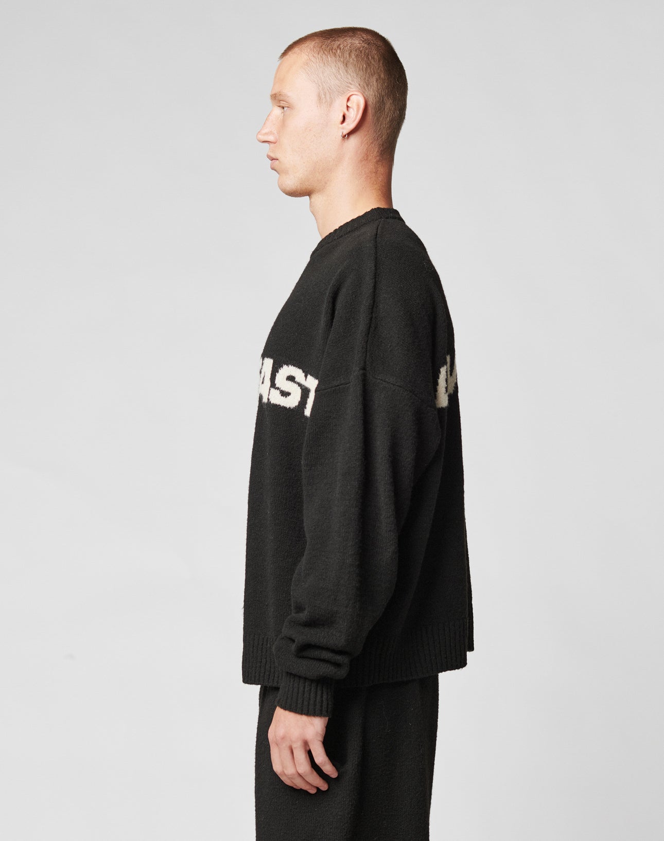 A man with short hair stands in profile against a plain background, wearing a black Live Fast Boucle Sweater by LIVE FAST DIE YOUNG, featuring partially visible white text, paired with matching black pants.