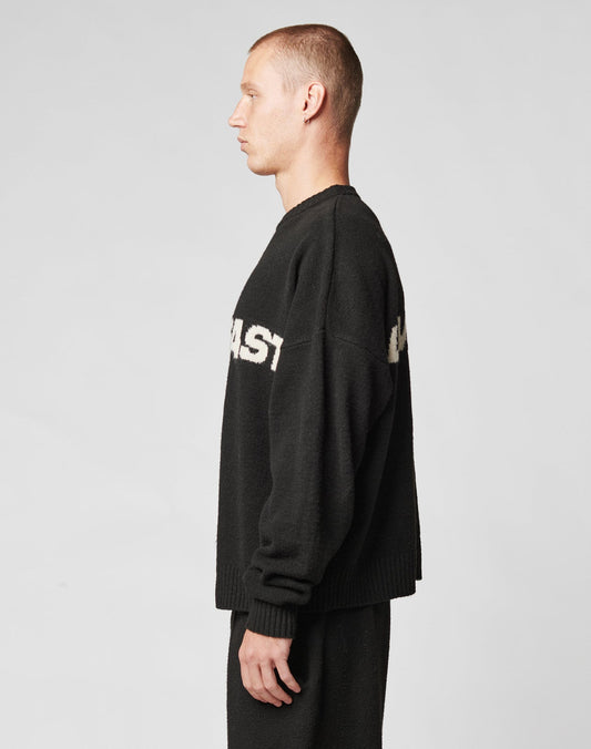 A man with short hair stands in profile against a plain background, wearing a black Live Fast Boucle Sweater by LIVE FAST DIE YOUNG, featuring partially visible white text, paired with matching black pants.