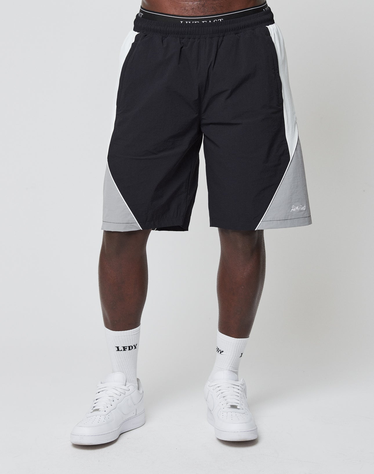 A person in LIVE FAST DIE YOUNG’s Basic Nylon Trackshorts (black, white & gray with elastisches Bündchen), white crew socks with “LFDY” text and Logo-Stick, and white sneakers—lower half pictured against a plain white background.