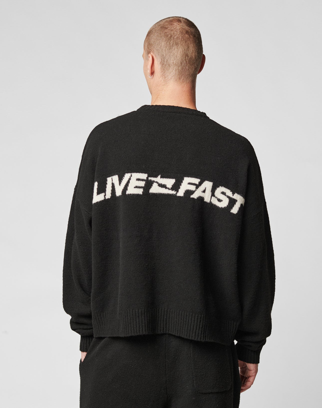 A person with short hair, seen from behind, wears the LIVE FAST DIE YOUNG Live Fast Boucle Sweater—an oversized black polyester knit featuring LIVE FAST in white capitals on the back. The background is plain light gray.