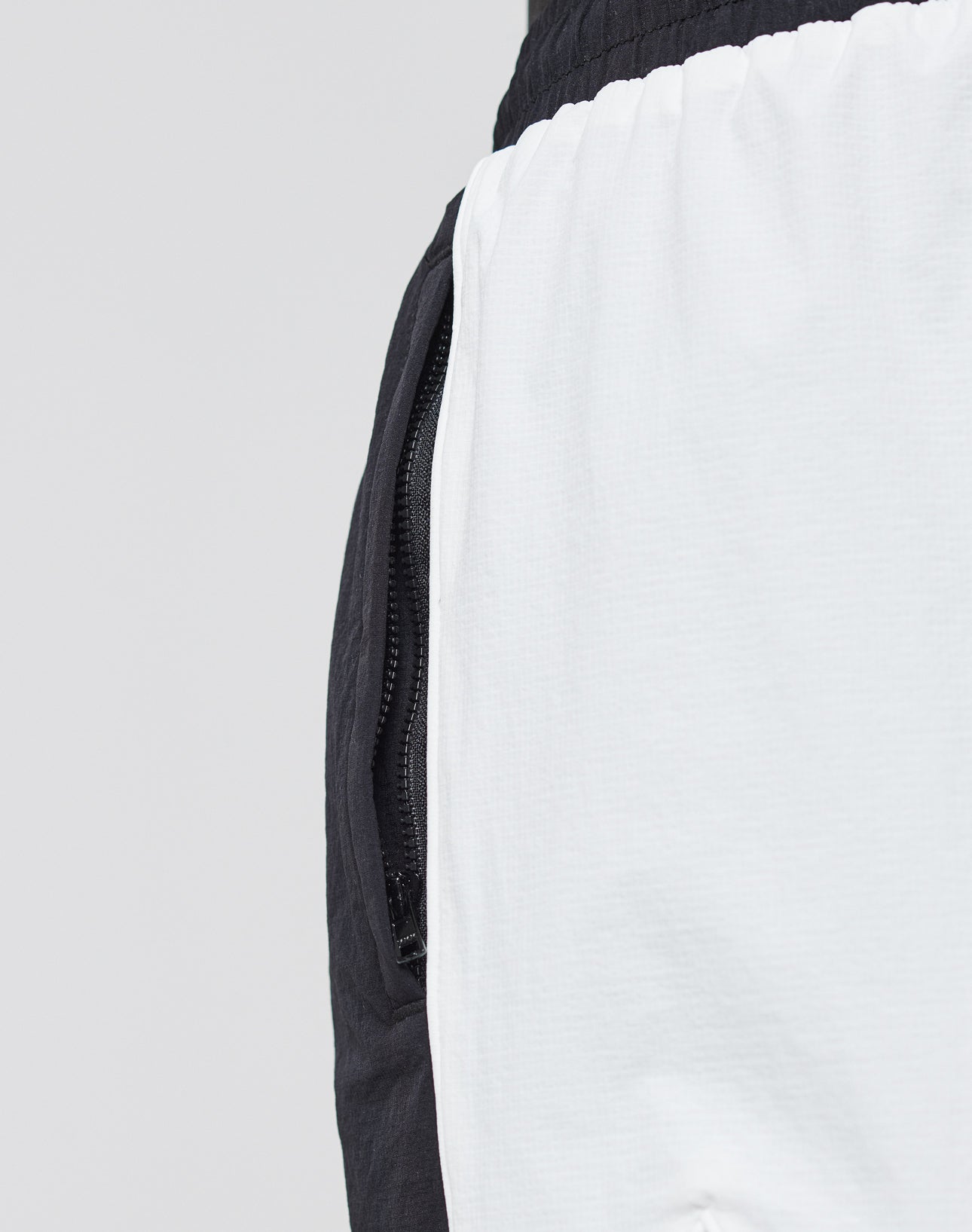 Close-up of a persons waist wearing LIVE FAST DIE YOUNG Basic Nylon Trackshorts in black and white color block. Features: elastic waistband, vertical side zip pocket, smooth nylon fabric. Plain light gray background.