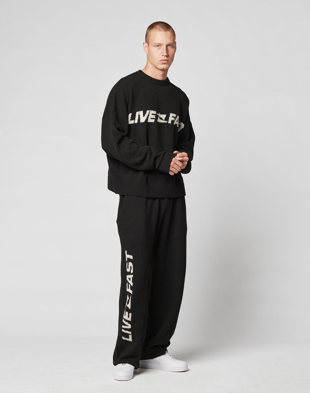 A man stands against a plain background in a wide fit black sweater and LIVE FAST DIE YOUNG’s Live Fast Boucle Pants, both with “LIVE FAST” in bold white letters, completed with white sneakers.