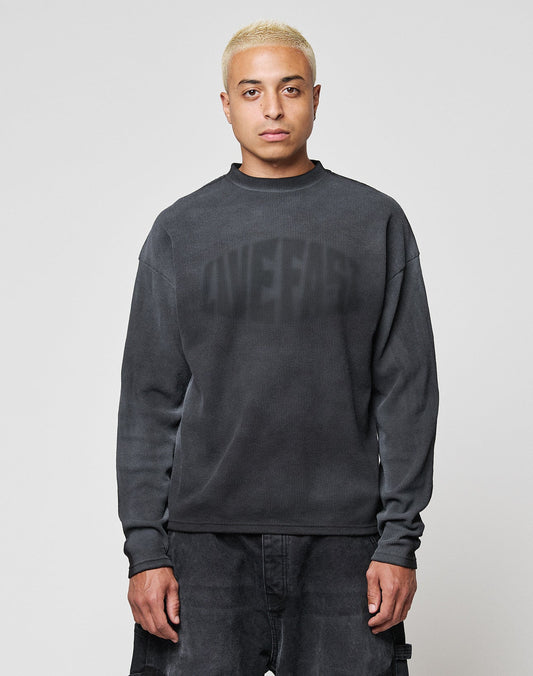 A person with short bleached blond hair wears the LIVE FAST DIE YOUNG LFDY Emblem Ghost Waffle Longsleeve, an oversized dark sweater with subtle lettering, and dark pants, standing facing forward against a plain light gray background.