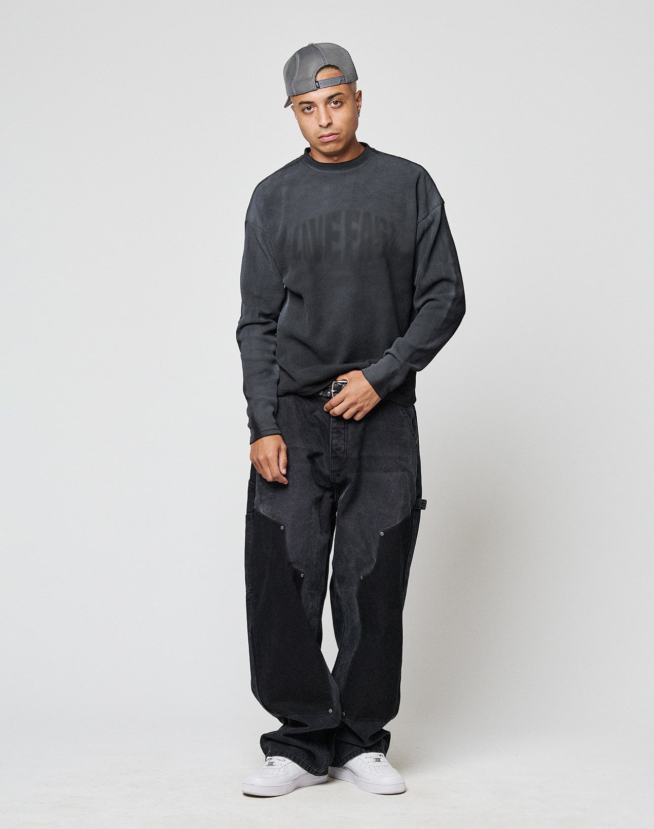 A person in a backwards gray cap, LIVE FAST DIE YOUNG LFDY Emblem Ghost Waffle Longsleeve, baggy black pants, and white sneakers stands against a light background with one hand in their pocket and a neutral expression.