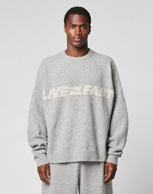 A person wears the LIVE FAST DIE YOUNG Live Fast Boucle Sweater in an oversized light gray fit with matching pants. The 100% polyester sweater displays blurred white LIVE FAST text across the chest. The background is plain and light-colored.| model