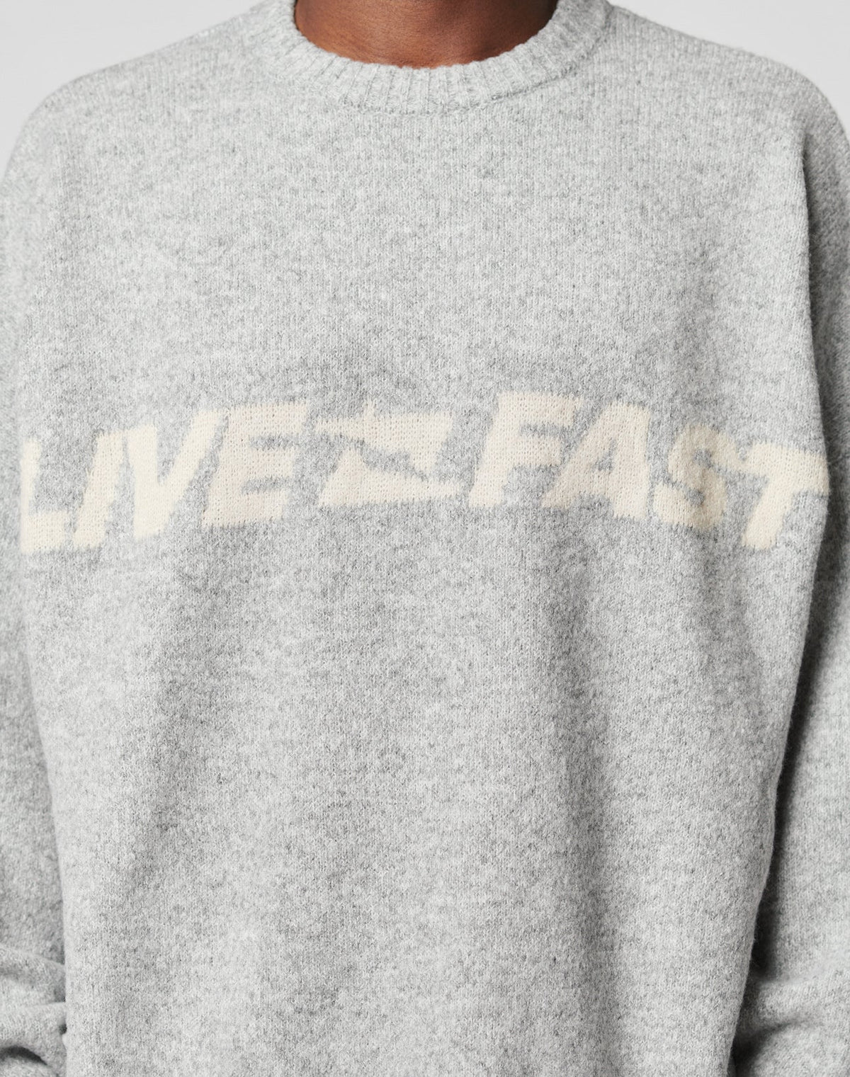 A person wears the LIVE FAST DIE YOUNG Live Fast Boucle Sweater, an oversize, light gray piece featuring LIVE FAST in off-white across the chest. Only their torso and neck are visible.