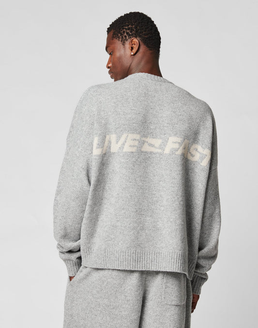 A person faces away, wearing an oversize light gray Live Fast Boucle Sweater and matching pants. The 100% polyester sweater by LIVE FAST DIE YOUNG displays LIVE FAST in bold white text across the back.