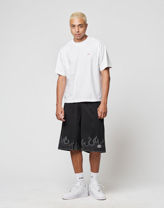 A person with short blond hair stands against a plain background, wearing a LIVE FAST DIE YOUNG LFDY Hinterhof Tee in white (100% Baumwolle), black shorts with a gray flame design, white socks, and white sneakers.